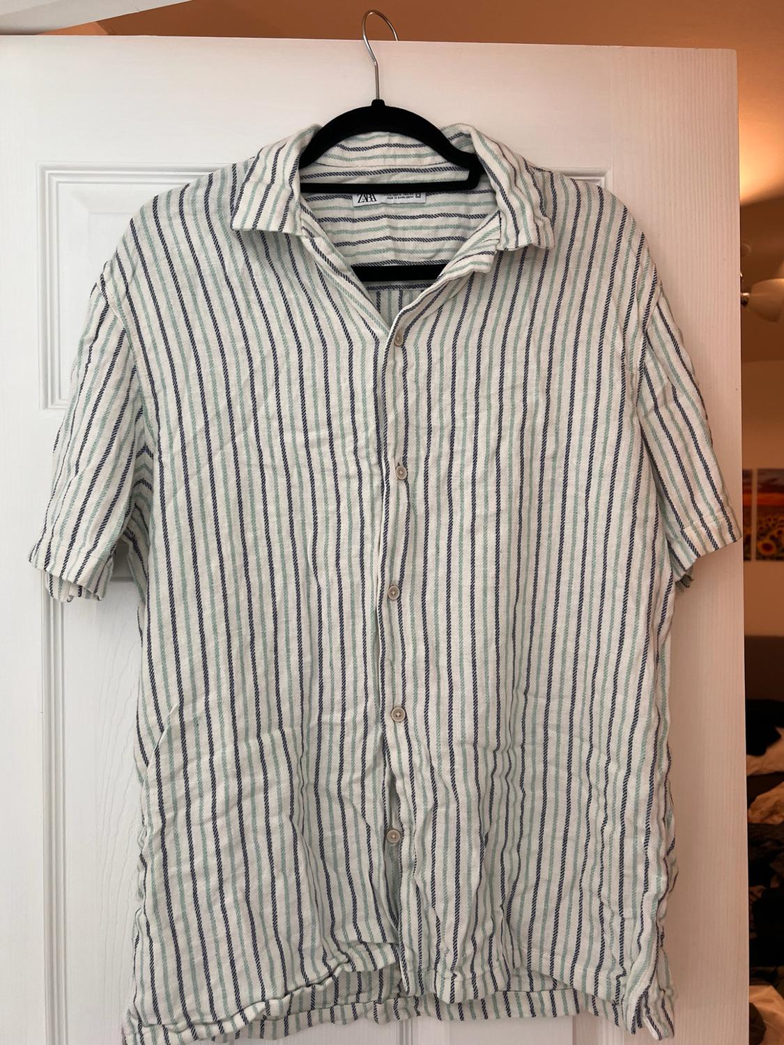 Short-sleeved shirt with vertical stripes hanging on a black hanger over a white door. The inner label reads ZARA.