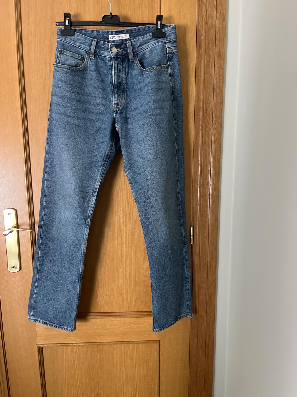 Blue jeans hanging on a wooden door with a black hanger. Visible label: "Zara".