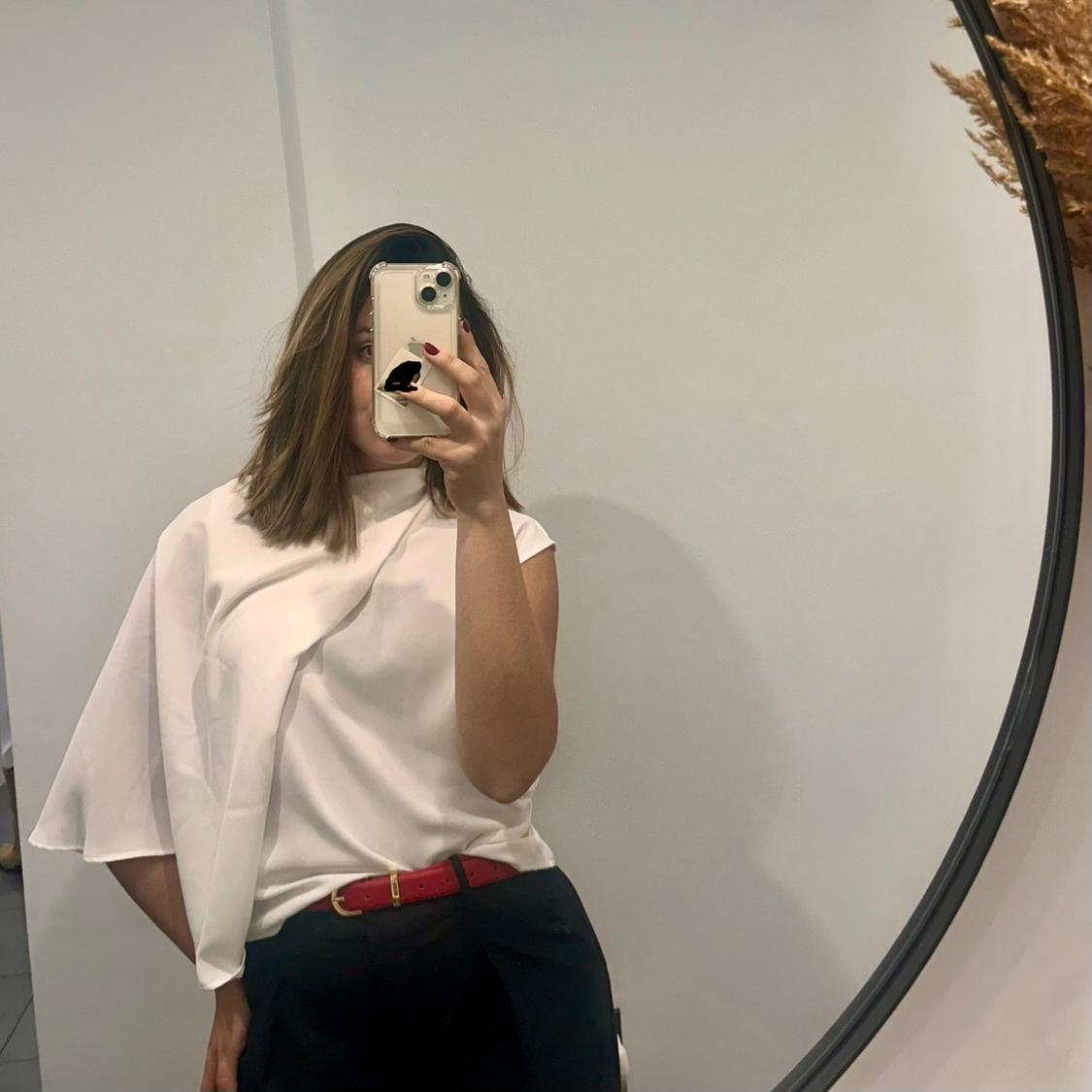 Woman in front of a mirror, wearing an asymmetrical white blouse, dark trousers, and a red belt. She is holding a mobile phone.