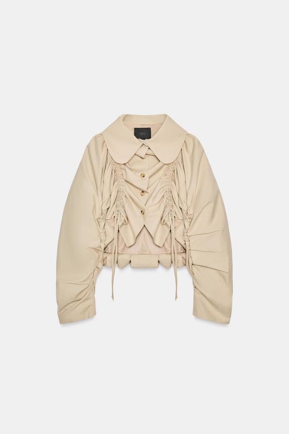 P�edn� pohled na b�ovou kr�tkou bundu s klopov�m l�mcem, dlouh�mi nab�ran�mi ruk�vy a p�edn�m zap�n�n�m na knofl�ky. Detail nab�ran�ch vol�nk� s v�z�n�m.