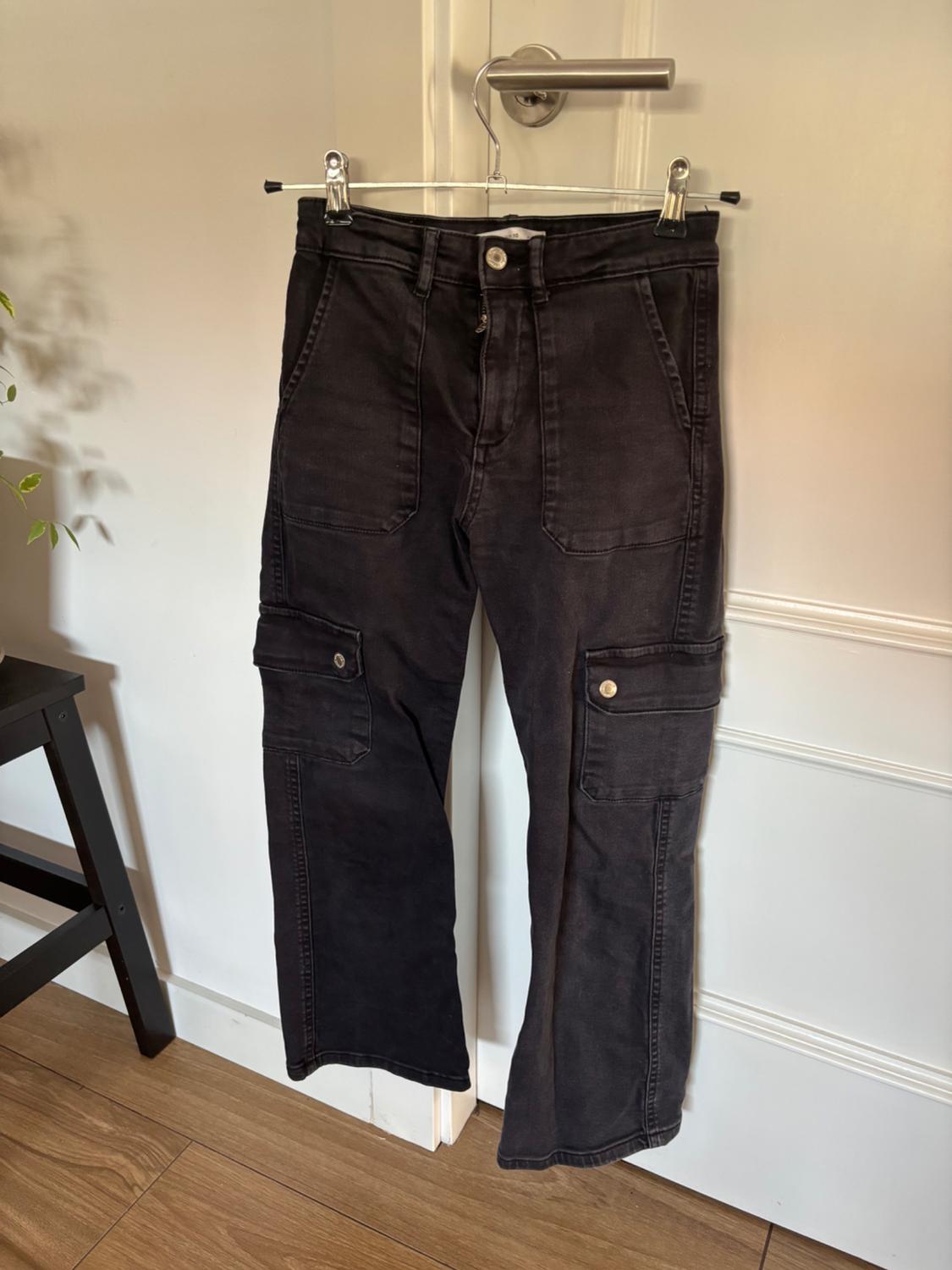 Black cargo trousers hanging on a hanger over a white door, with large pockets and a metal button. Wooden floor.