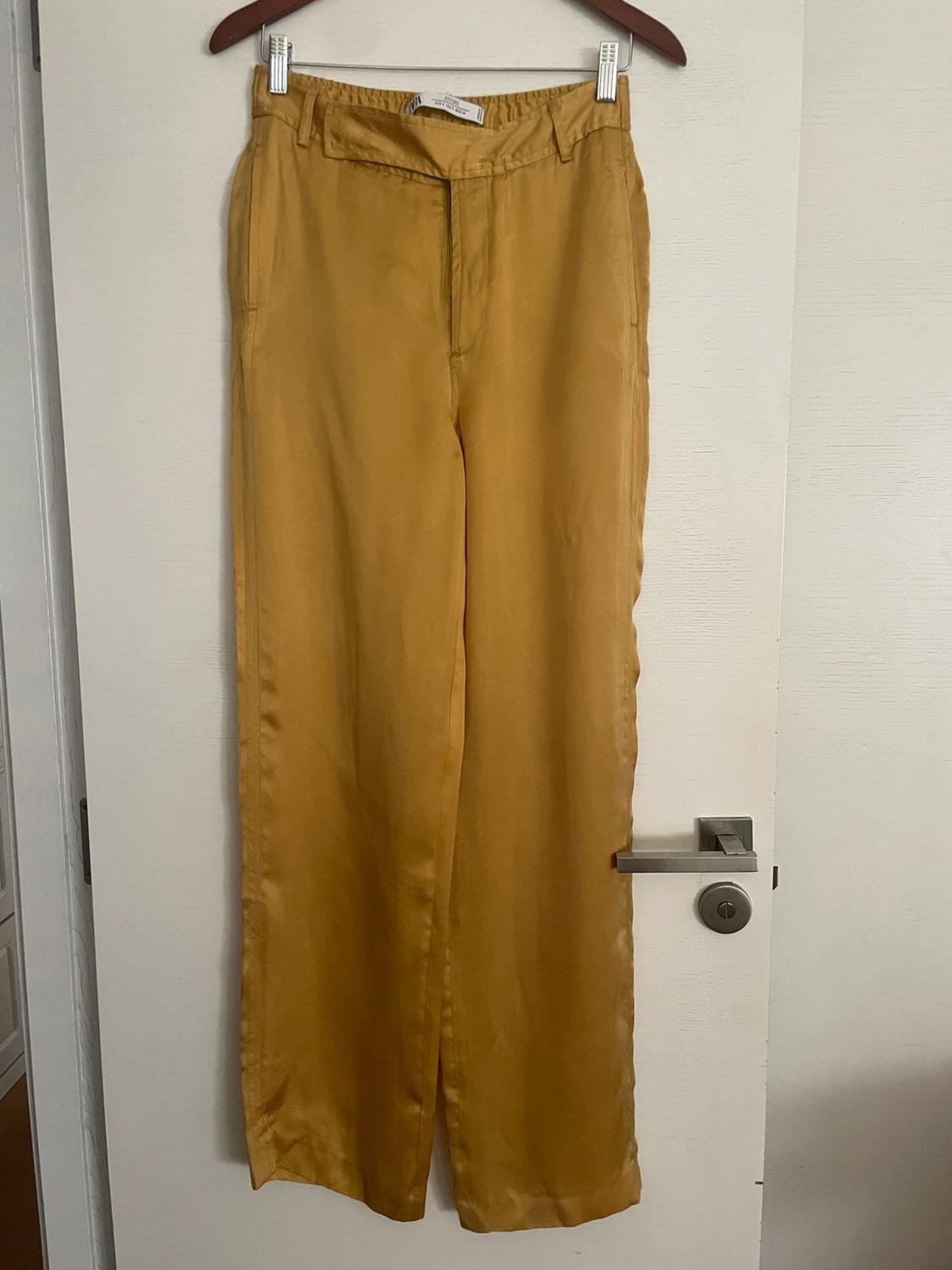 Mustard-coloured satin trousers, hanging on a wooden hanger on a white door with a metal handle.