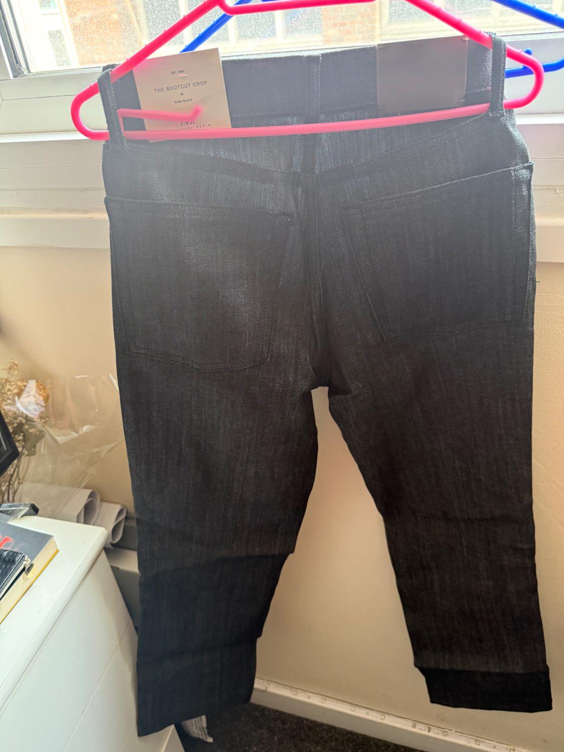 Rear view of black jeans on a pink hanger in front of a window. Text: THE BOOTCUT CROP IN DUSK BLACK ZARA SIGNATURE DENIM.