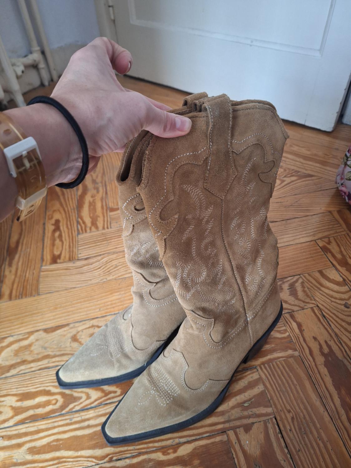 Hand holding brown suede cowboy boots with embroidered design, on light wooden floor. Background with white door.