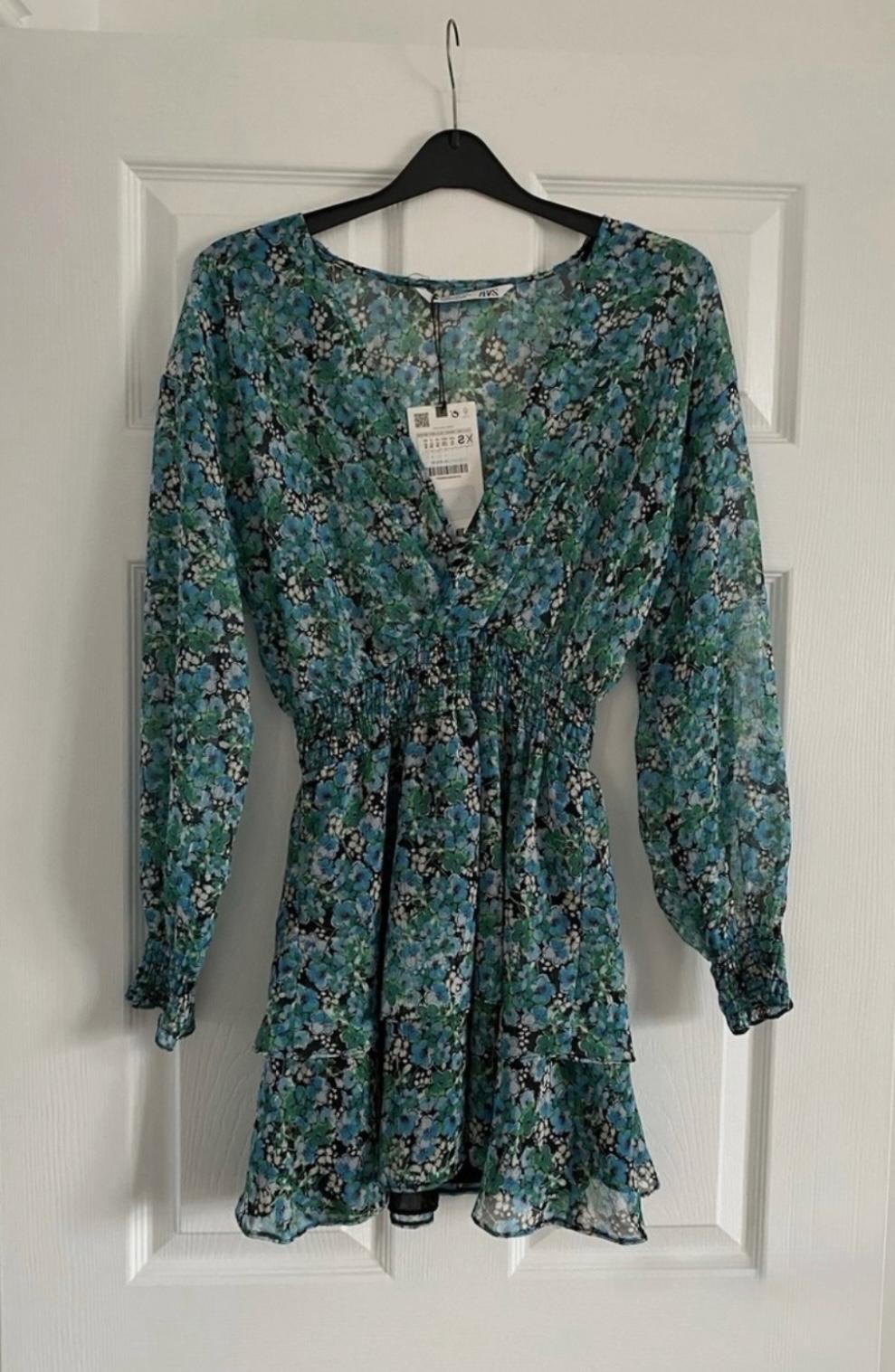 Blue and green floral dress with long sleeves, hanging on a black hanger over a white door. Label visible at the neckline.