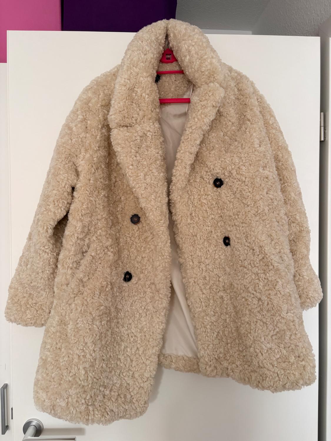 Beige teddy coat hanging on a white door, with black buttons and a pink hanger. Purple and pink paneled background.