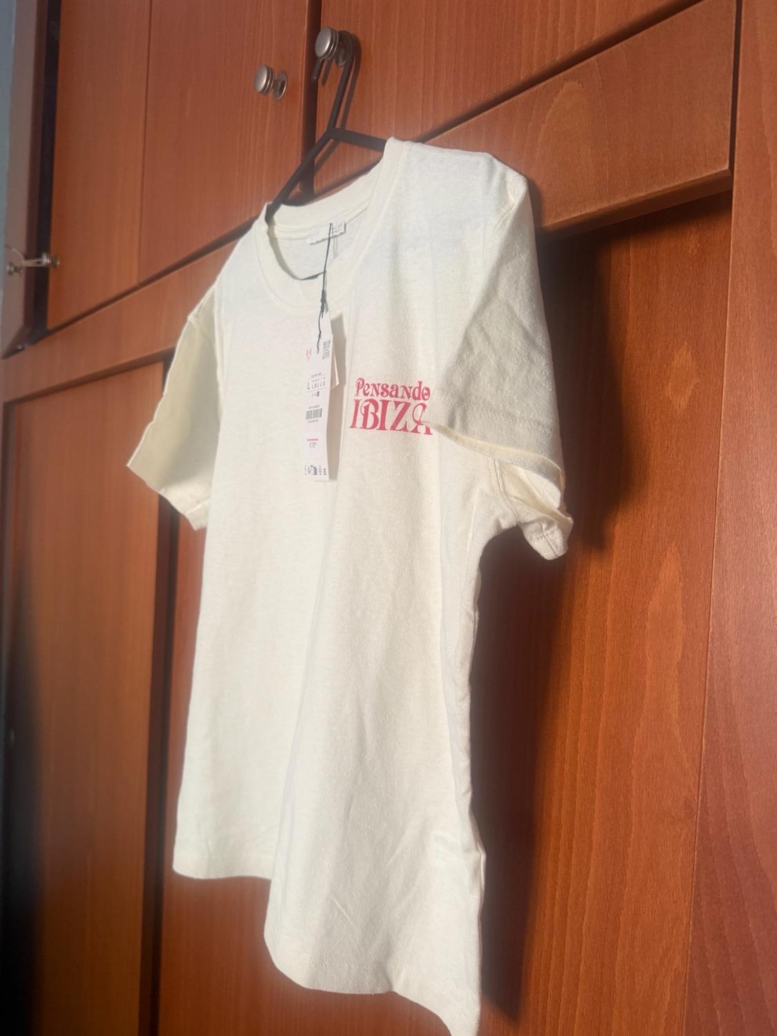 White short-sleeved T-shirt with a label, hanging in a wooden wardrobe. The red text “Thinking IBIZA” is printed on the chest.