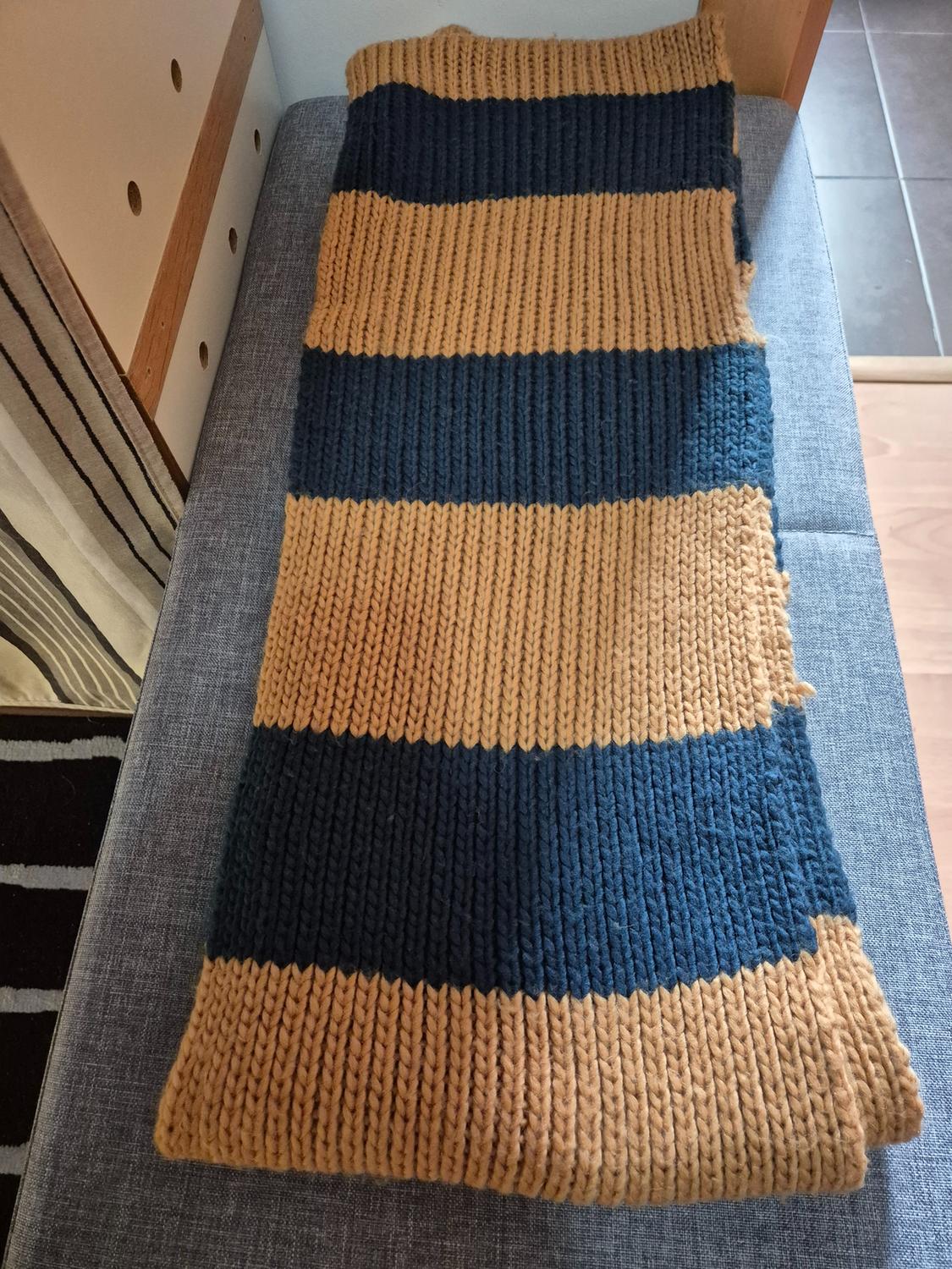 Chunky knit scarf with horizontal dark blue and mustard stripes, placed on a grey upholstered bench.