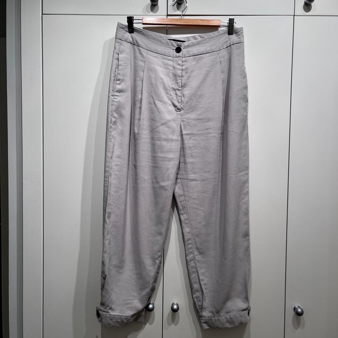 Grey trousers hanging on a wooden hanger in front of a white wardrobe. Featuring pleats and a rolled hem.