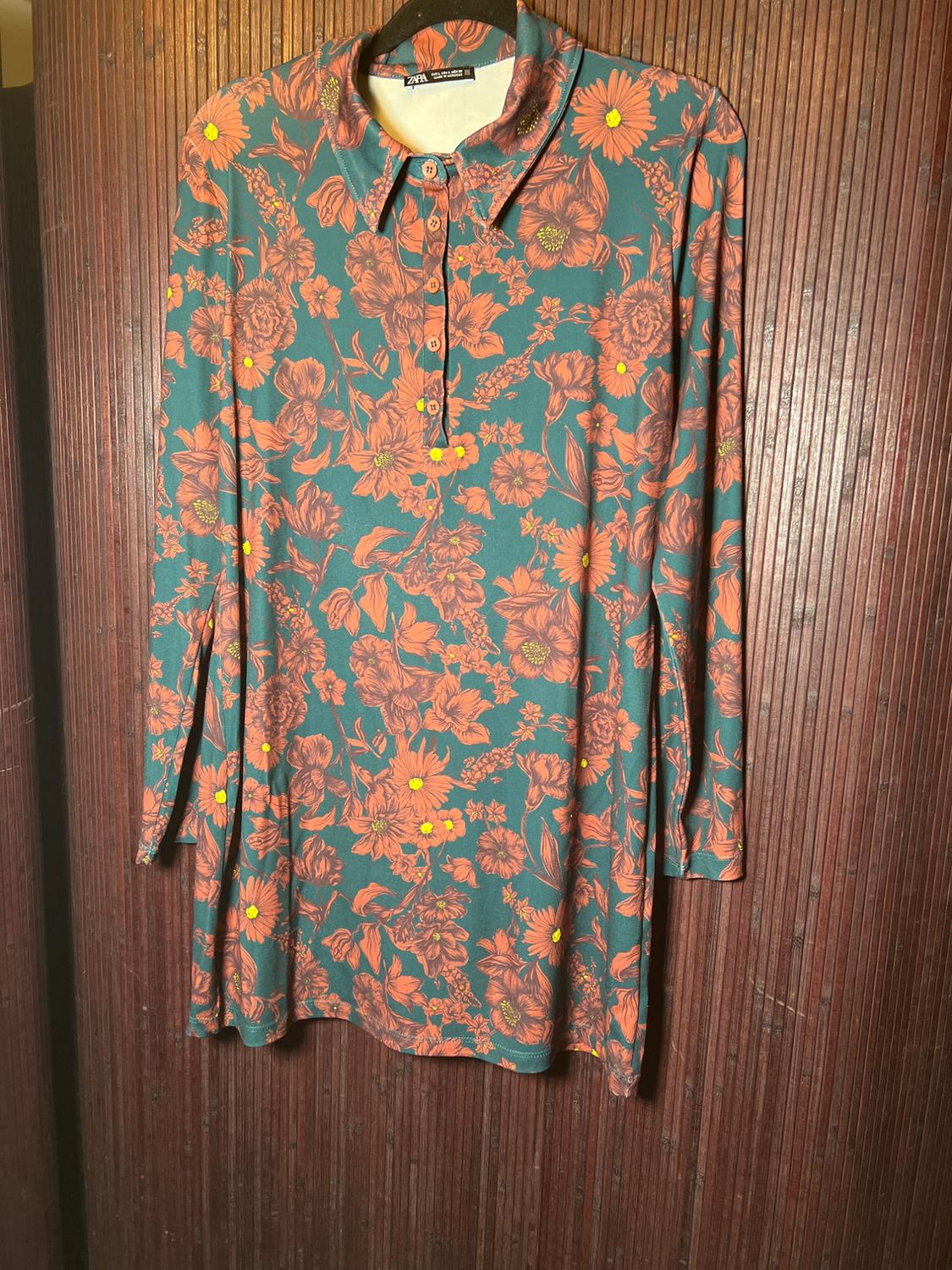 Long-sleeved dress with a floral print in orange and yellow tones on a green background, hanging on a wooden hanger.