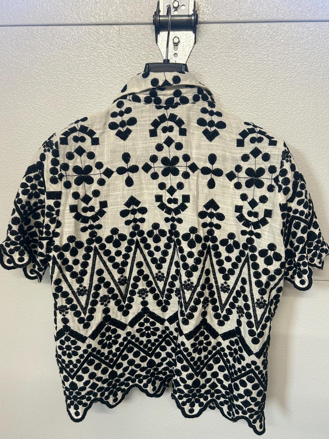 White short-sleeved shirt seen from the back, with geometric and floral black embroidery, hanging on a hanger against a light background. Scalloped hem.