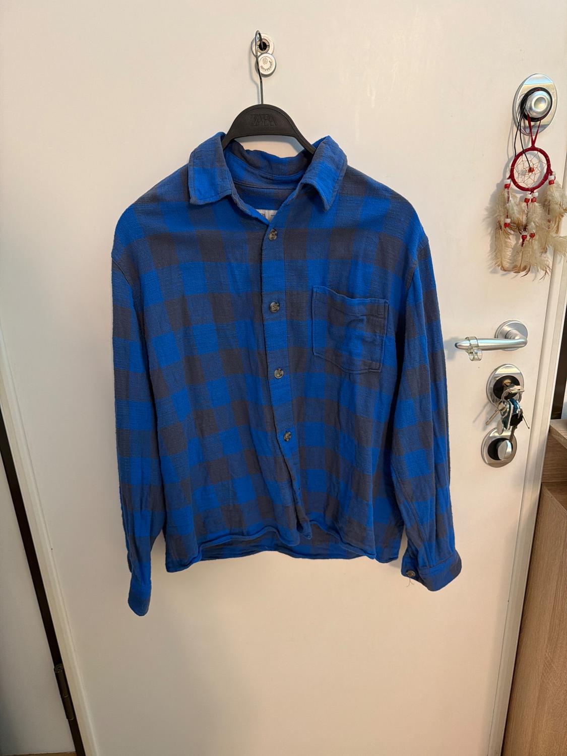 Blue and grey checked shirt with pocket, on ZARA hanger hanging on a white door with keys and dreamcatcher. Casual style.