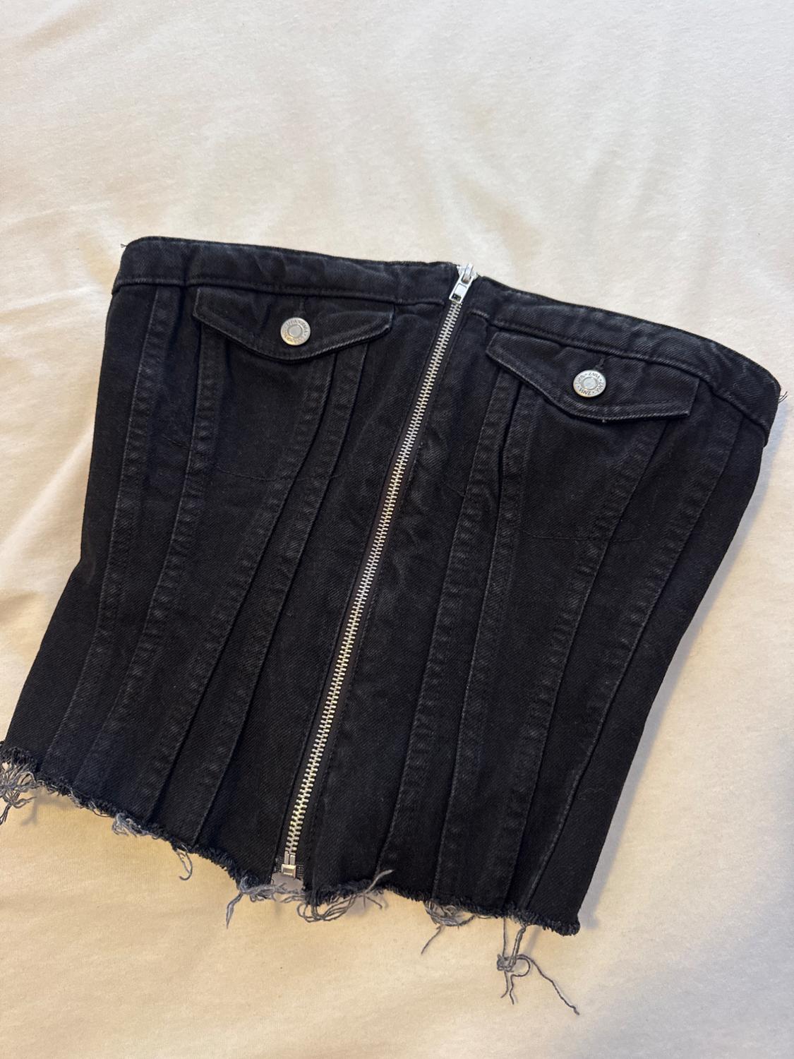 Black denim bandeau top with a front zip, pockets and a frayed hem, set against a light fabric background.
