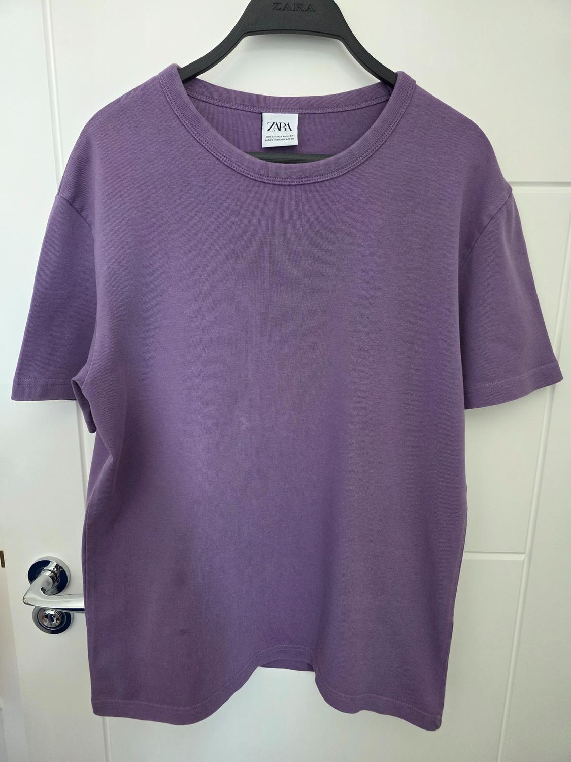 Purple short-sleeved T-shirt hanging on a black hanger in front of a white door. Visible text: ZARA.