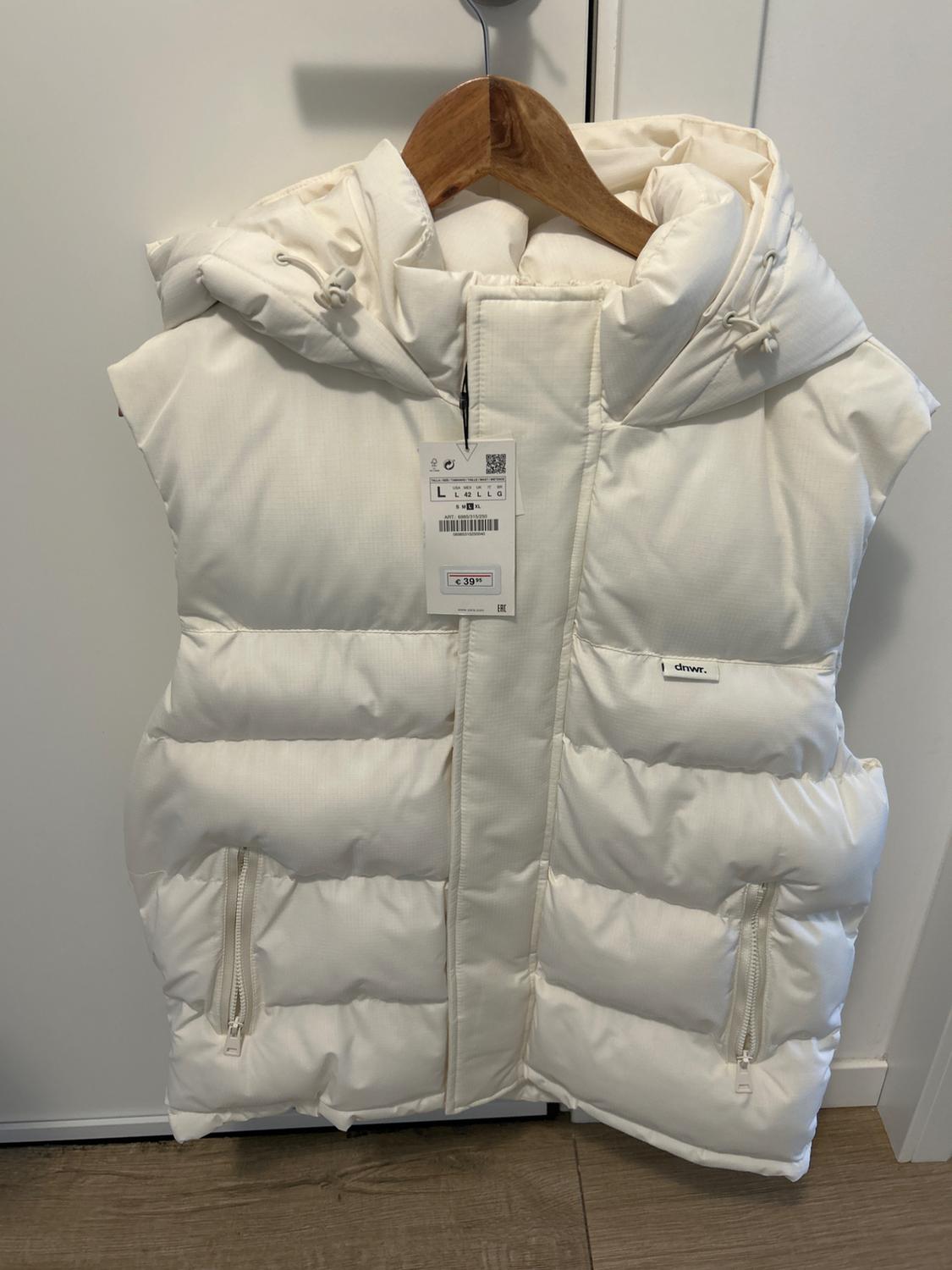 White padded gilet with hood and zip fastening, hanging on a wooden hanger. Visible tag with price of €39.