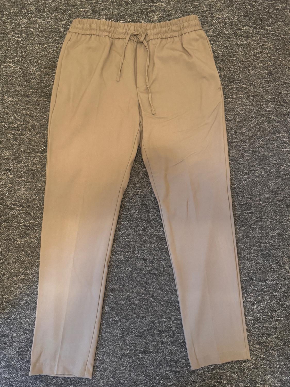 Beige trousers with an elasticated drawstring waist, laid on a textured grey surface.