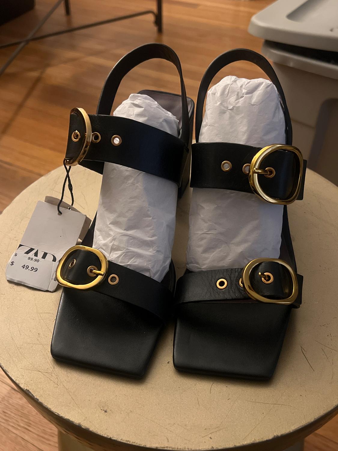 Black block heel sandals with gold buckles on a wooden table. Label shows price of £49.99.
