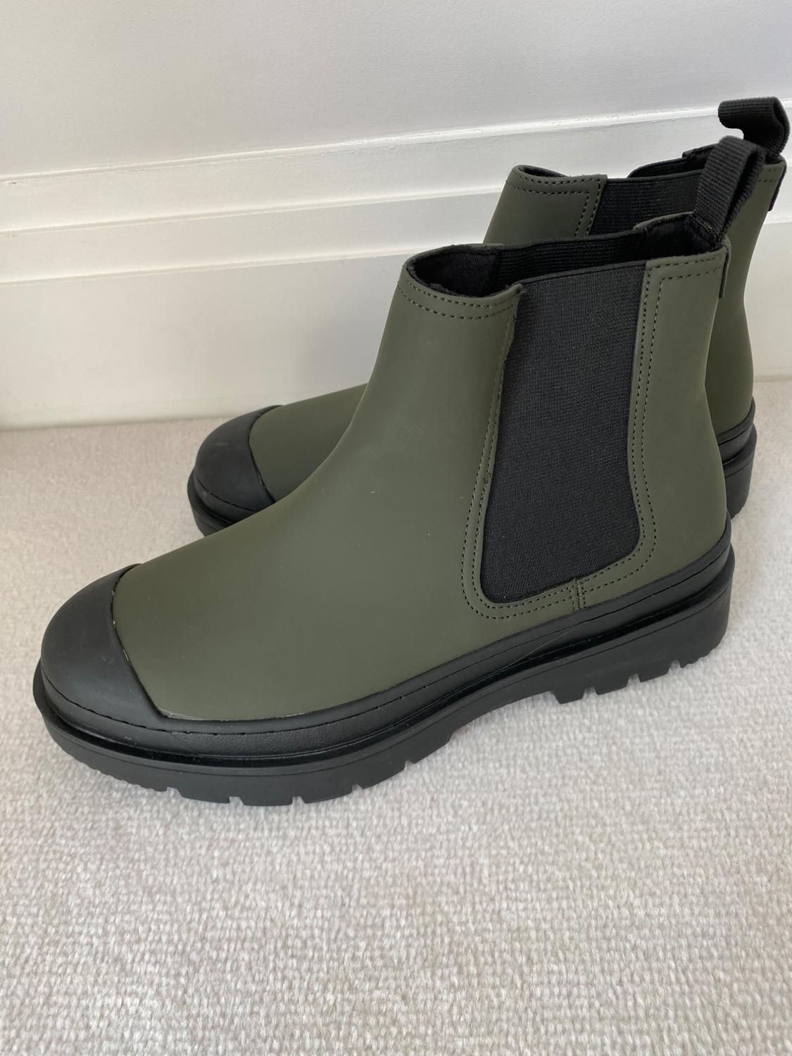 Green boots with black toe cap, black elastic side panels and chunky black sole, on beige carpet with white wall in the background.