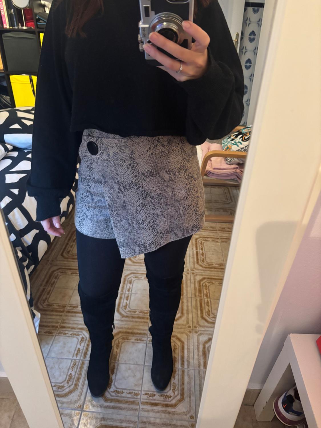 Woman in mirror wearing a black jumper, grey skirt with snake print, leggings, and black boots. Room background with tiled floor.