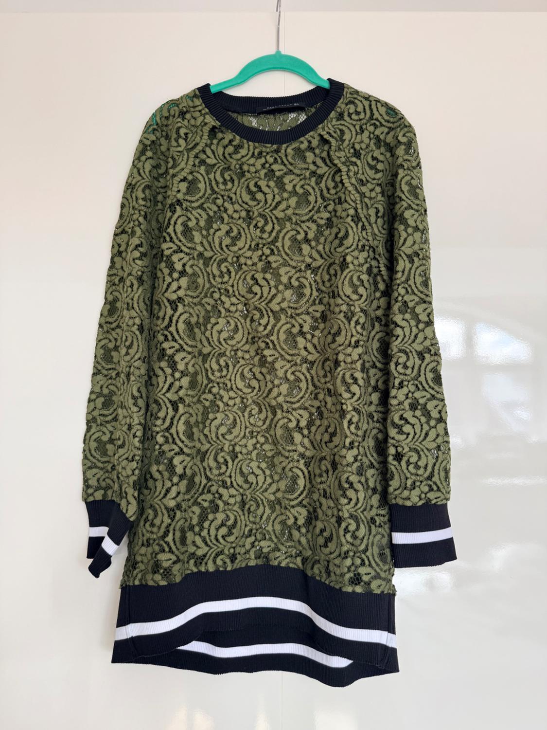 Olive-green lace long top with a black collar, cuffs and hem featuring white stripes, hanging on a green hanger against a white background.