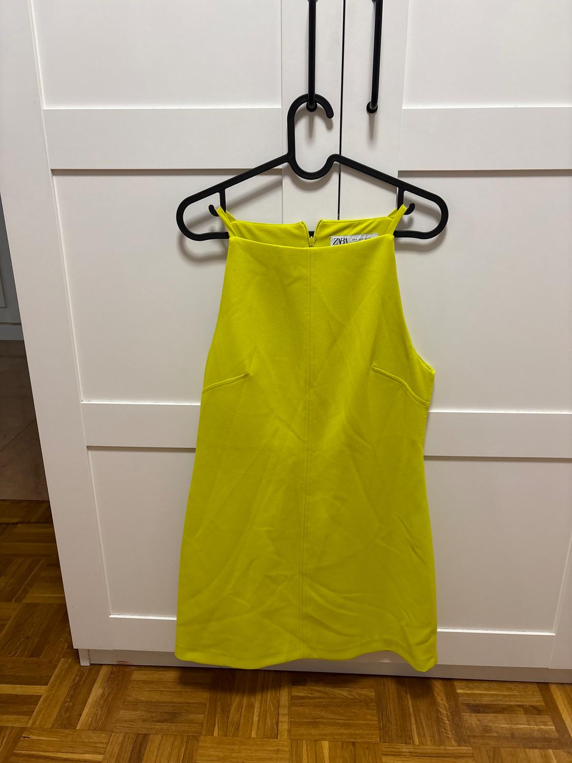 Short yellow sleeveless dress with ZARA text, hanging on a black hanger in a white wardrobe. Wooden floor at the bottom.