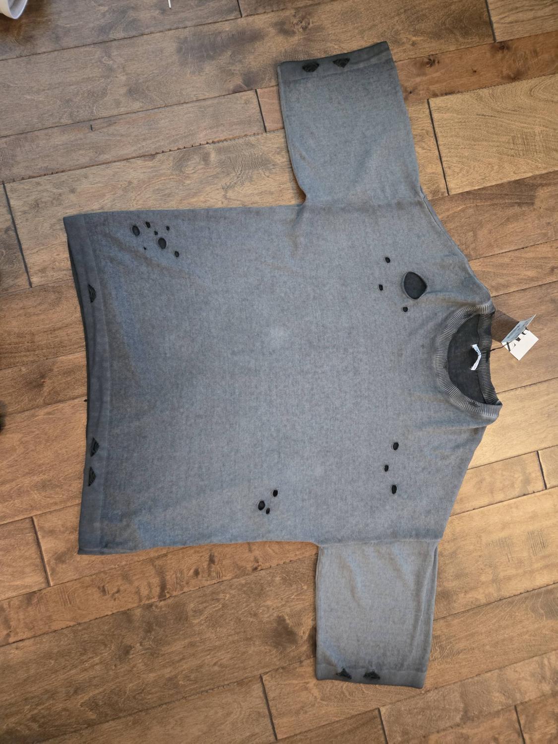 Gray short-sleeve T-shirt with a distressed effect and decorative holes, placed on a wooden floor.