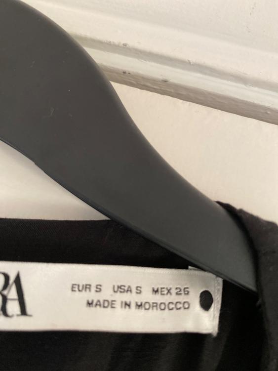 Clothing label on a black hanger, visible text: "EUR S USA S MEX 26 MADE IN MOROCCO". Black and white background.