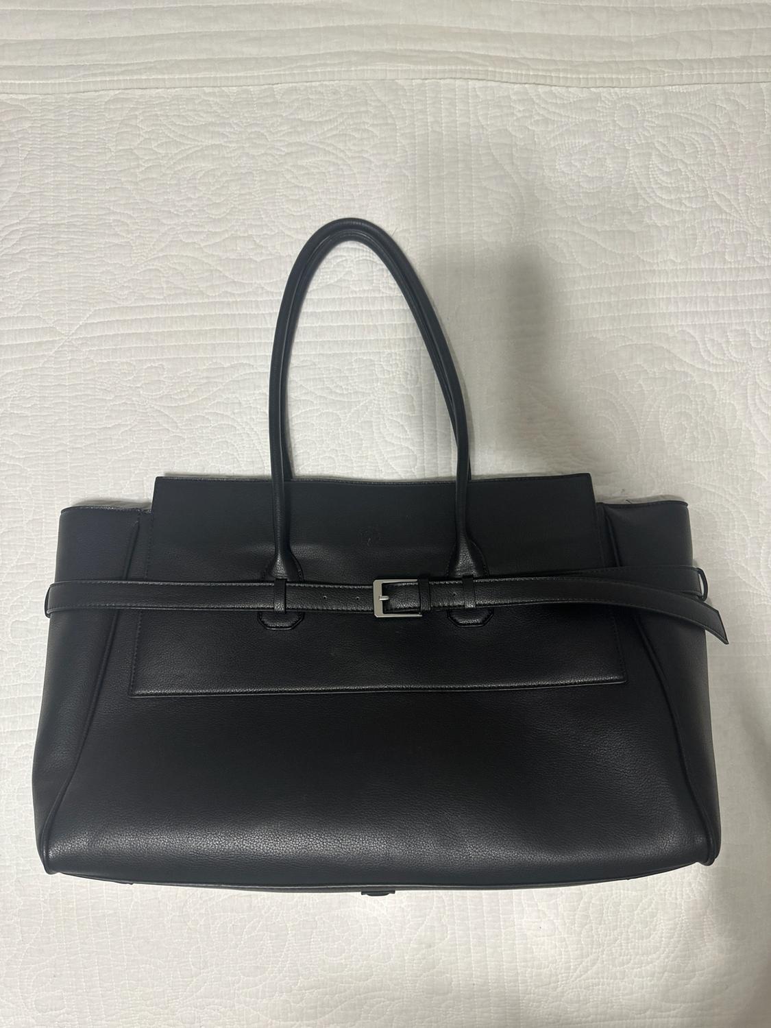 Black handbag with handles and a belted detail featuring a silver buckle, set against a white quilted surface.