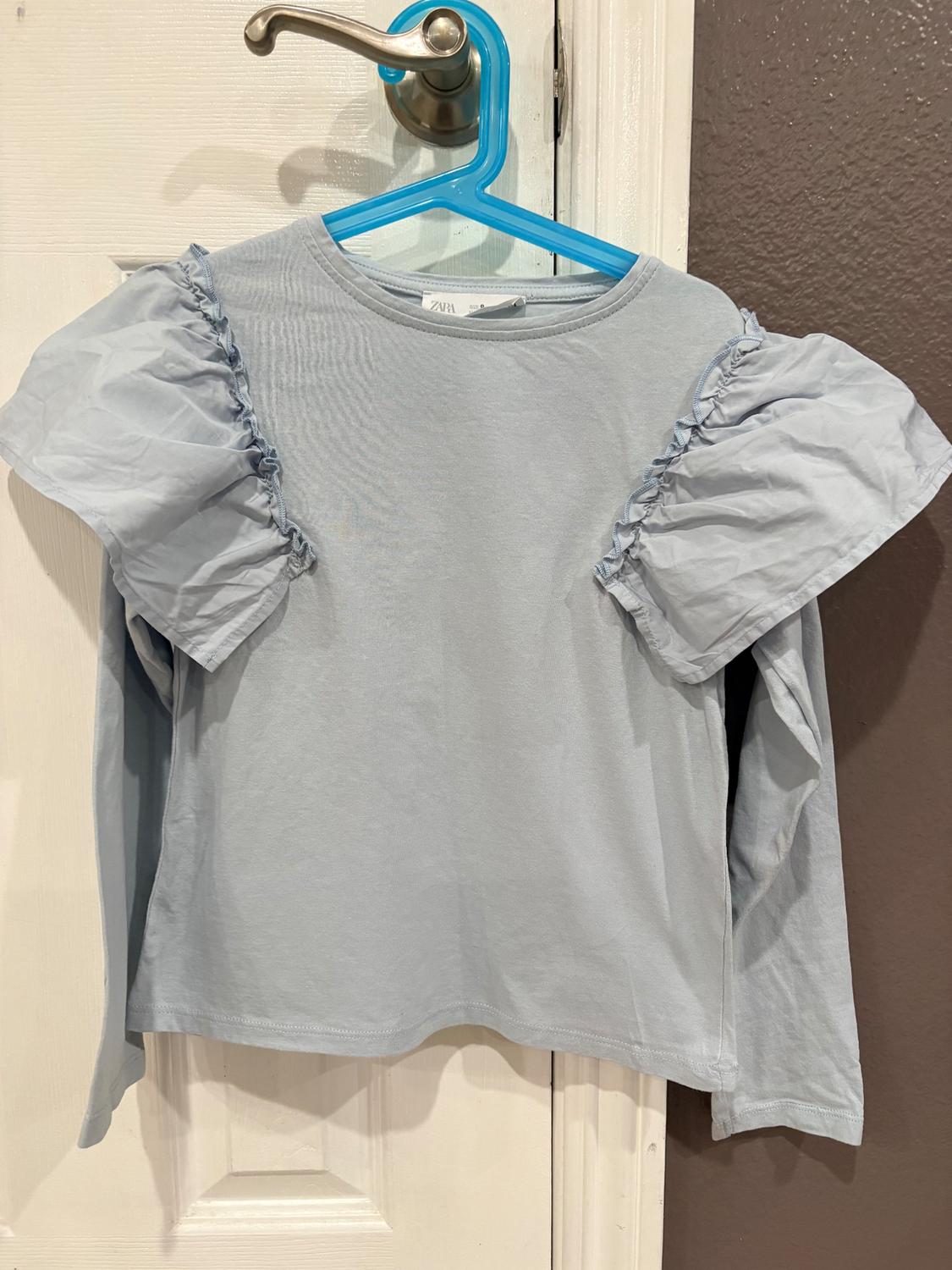 Light blue blouse with long sleeves and ruffles on the shoulders, hanging on a blue hanger over a white door.