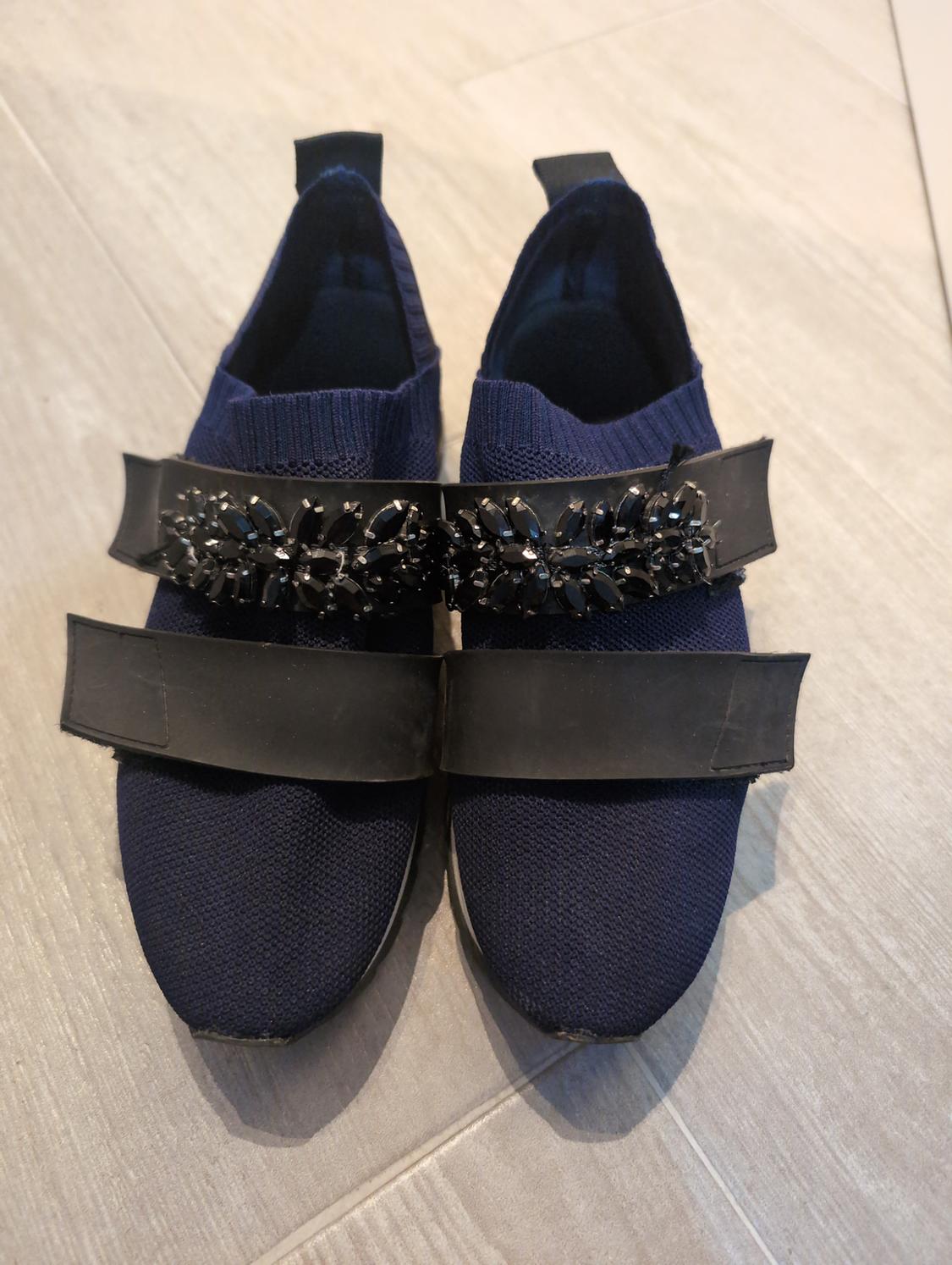 Navy knitted slippers with two black front straps, one plain and one with black rhinestones, on a light wooden floor.