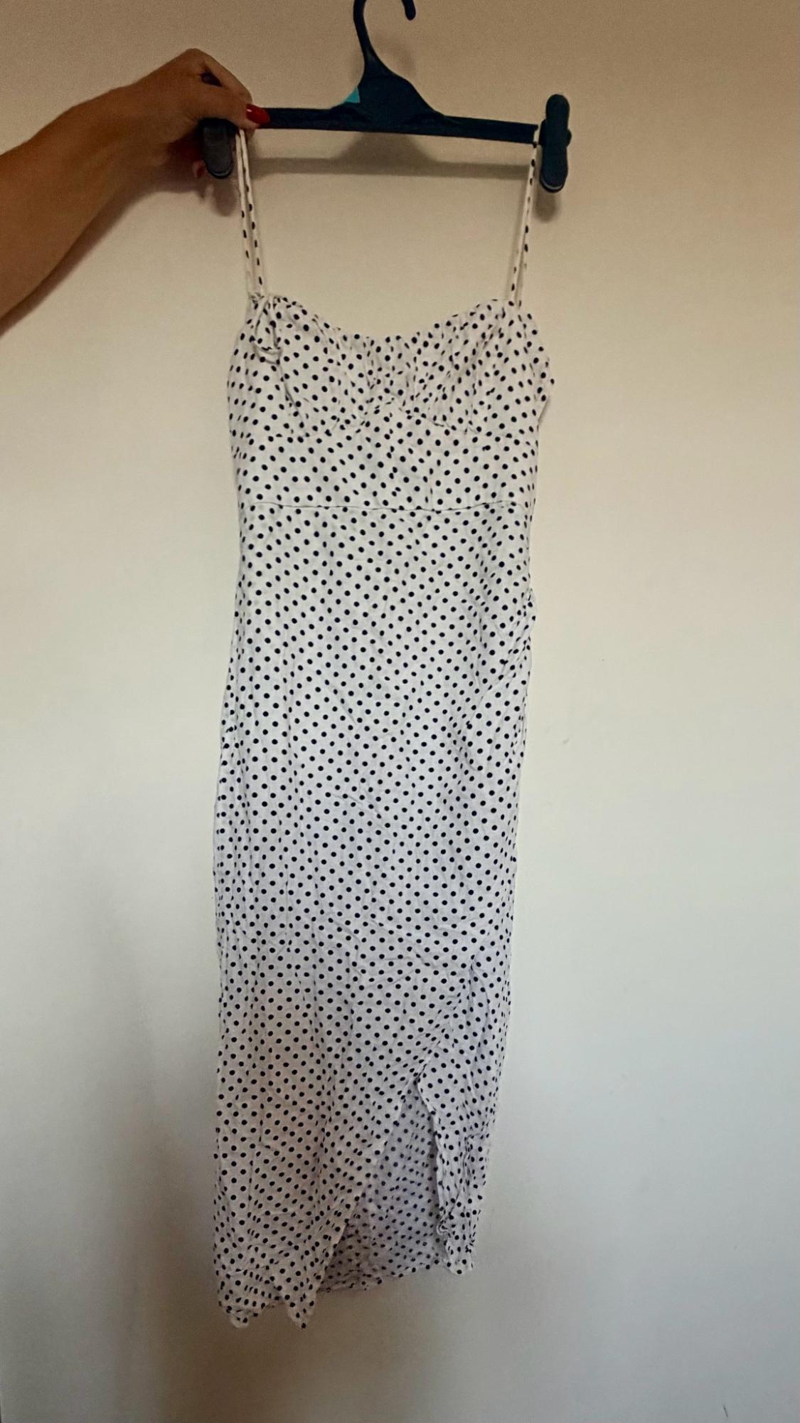 Hand holding a hanger with a white dress featuring black polka dots, spaghetti straps, and a front slit in front of a white wall.