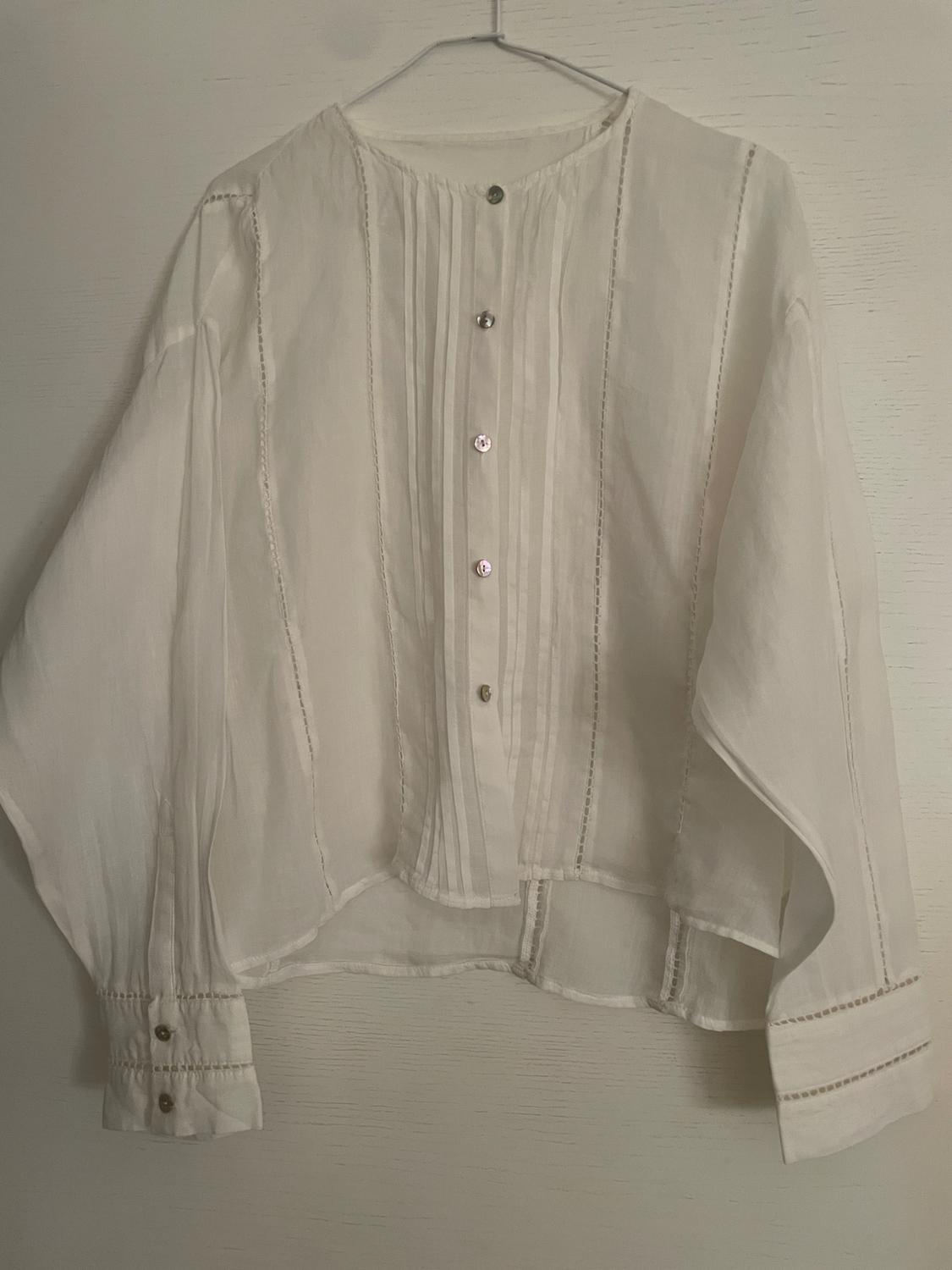White long-sleeved blouse on a hanger against a light background. Features front pleats, button fastening and openwork details on the seams.
