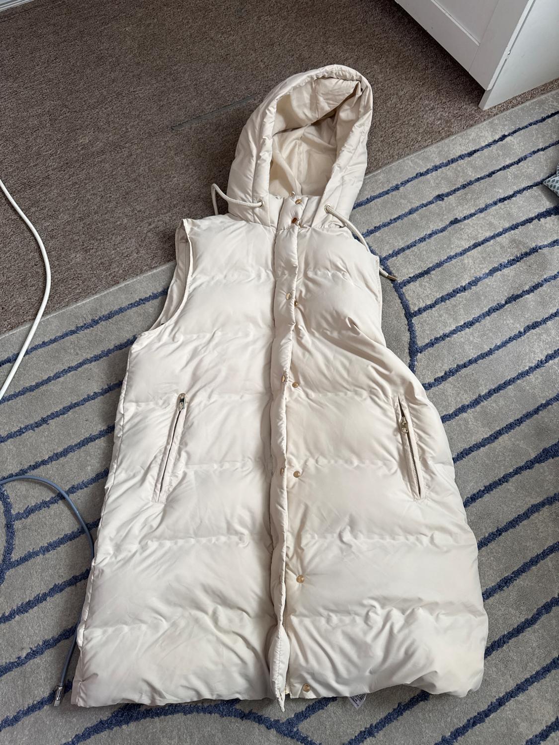 Cream long padded gilet with a hood and buttons, laid out on a grey rug with blue stripes in a relaxed setting.