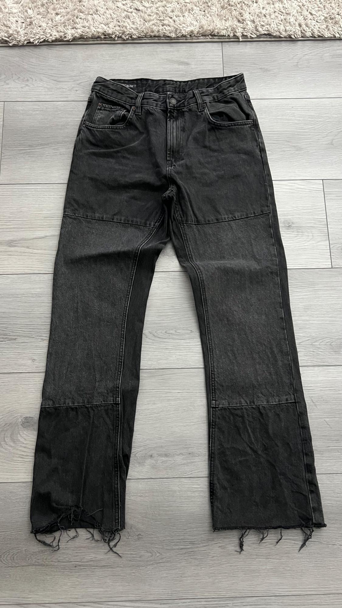 Black denim pants with horizontal stitching and frayed hems, laid flat on a light wooden floor. Casual and relaxed style.