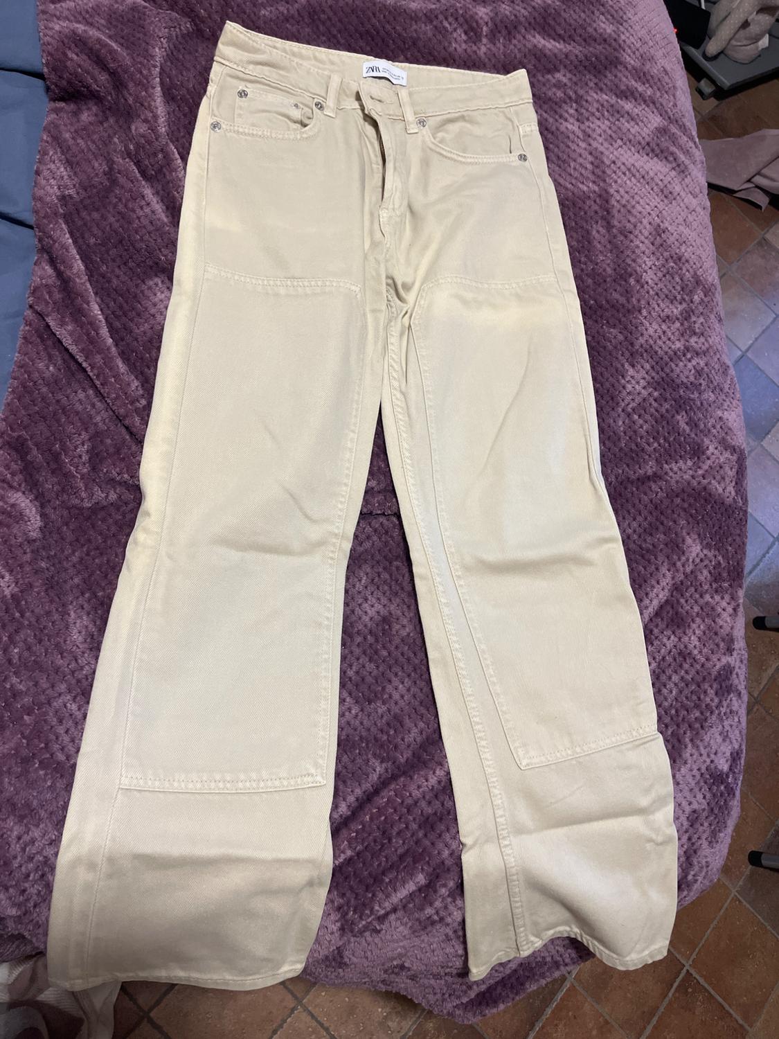 Beige straight-cut trousers on a textured purple blanket. Visible waist label. Brown tiled background.