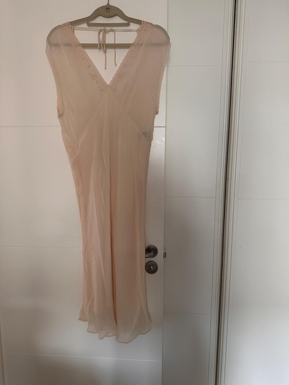 Μακρύ φόρεμα από γάζα σε nude χρώμα κρεμασμένο σε μια κρεμάστρα πάνω από μια λευκή πόρτα, με λεπτομέρειες δαντέλας στους ώμους.