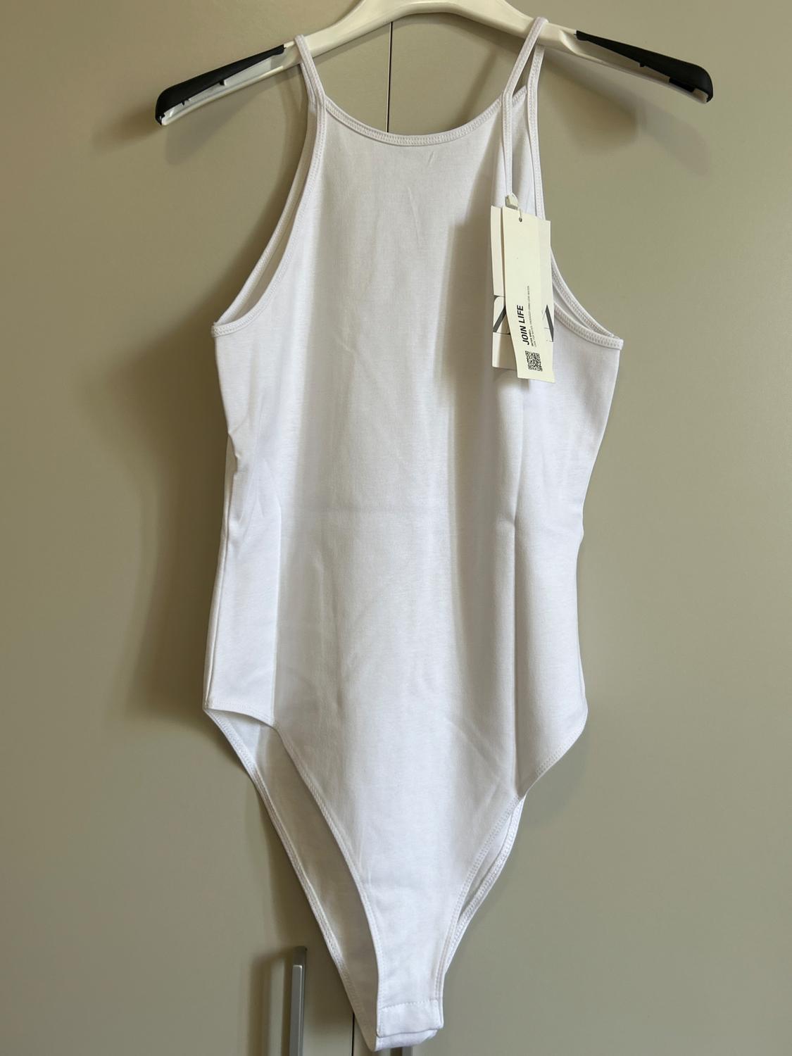White high-neck, strappy bodysuit hanging on a hanger in front of a light-coloured wardrobe. Includes a tag with the text "JOIN LIFE".
