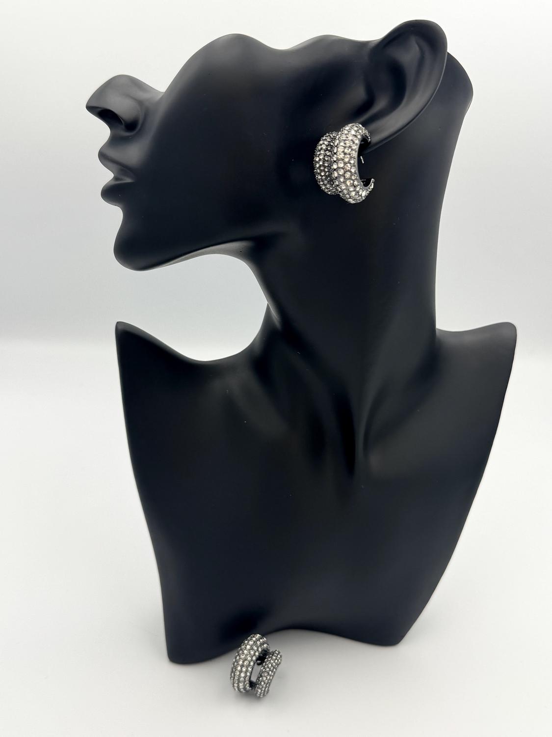 Black mannequin with a shiny silver earring on the ear. Another similar earring is displayed at the base of the mannequin. White background.