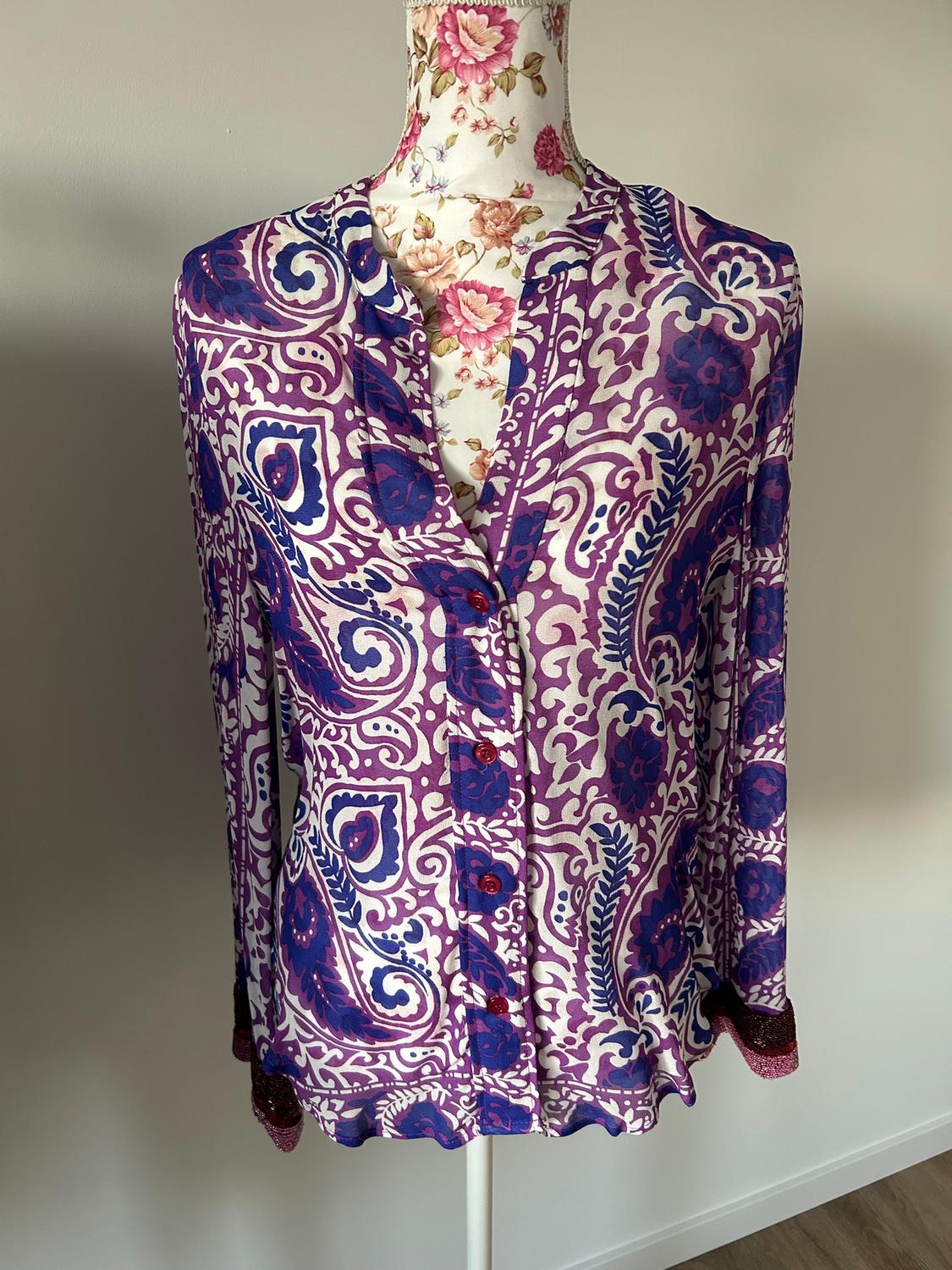 Long-sleeved blouse with floral print in purple and blue tones on a mannequin with floral fabric against a neutral background.