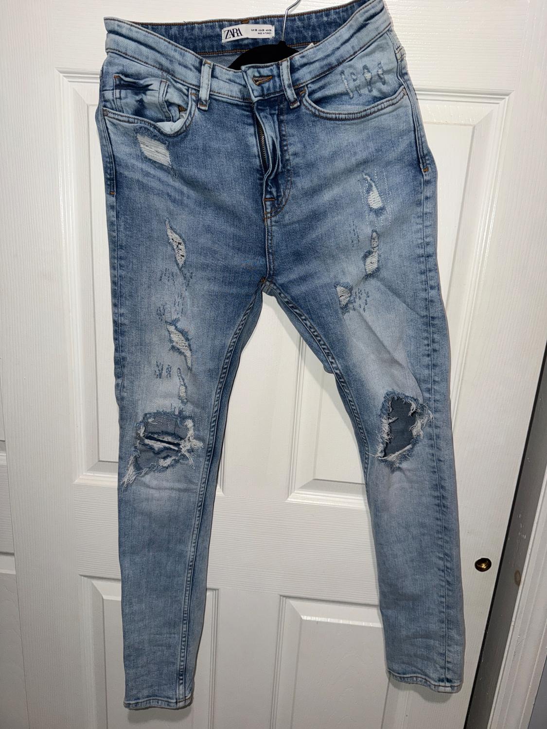 Worn blue jeans hanging on a white door. The label says "Zara". Visible tears on the legs.