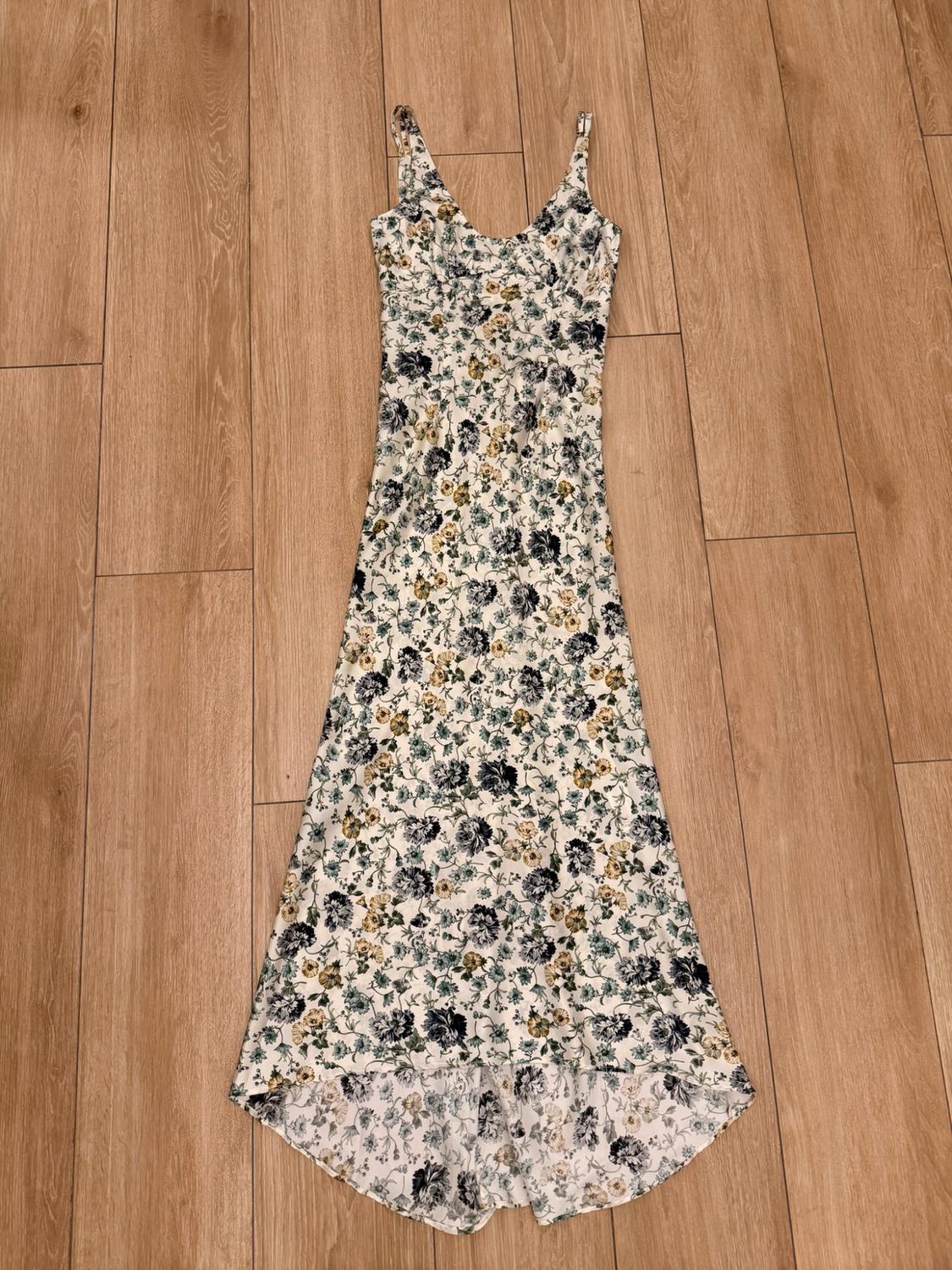 Long strappy dress with a blue and yellow floral print on a white background, V-neck and asymmetrical hem, on a wooden floor.