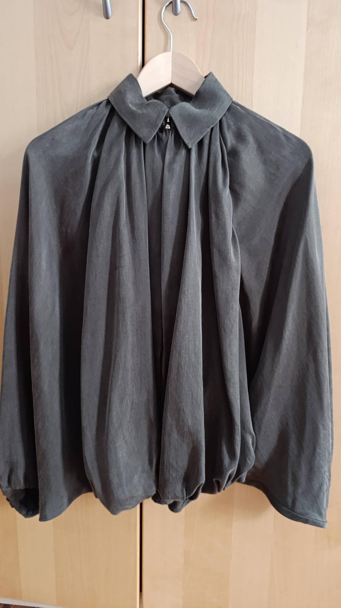 Dark grey relaxed-fit blouse with a collar, zip fastening and gathered hem, hanging on a hanger against a light wood background.