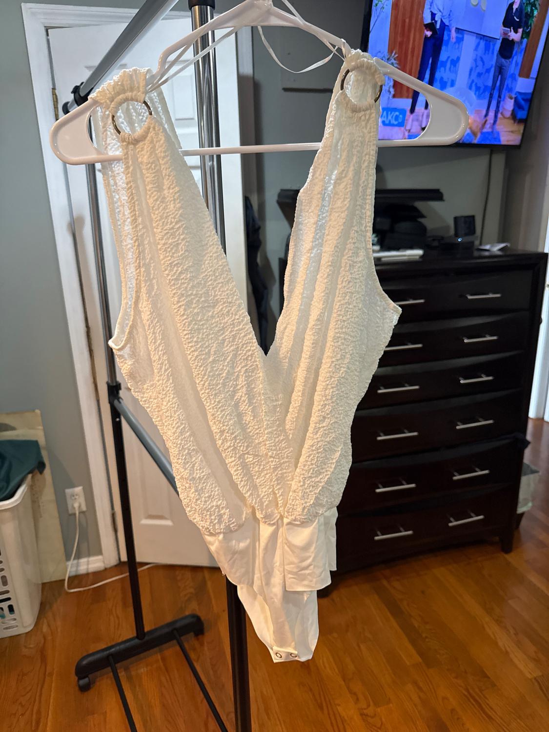Textured white bodysuit hanging on a hanger in a room with a wooden floor and a TV on in the background.