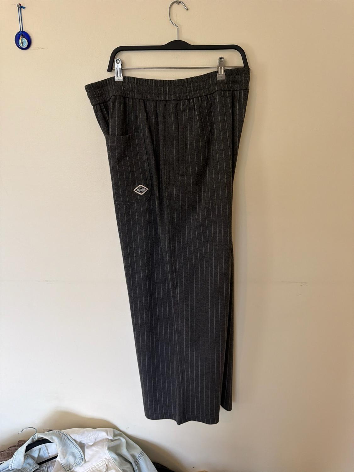 Dark grey trousers with white stripes, elasticated waist and side pocket, hanging on a hanger against a light wall. Casual style.