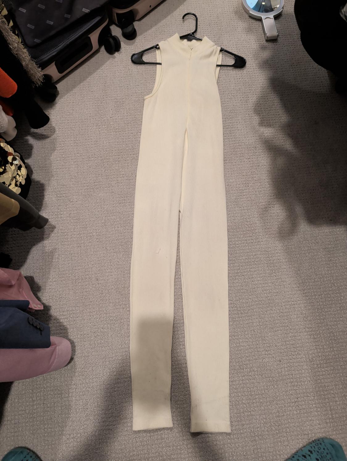 Cream sleeveless jumpsuit hanging on a hanger over a beige carpet, surrounded by clothes and suitcases in a wardrobe.