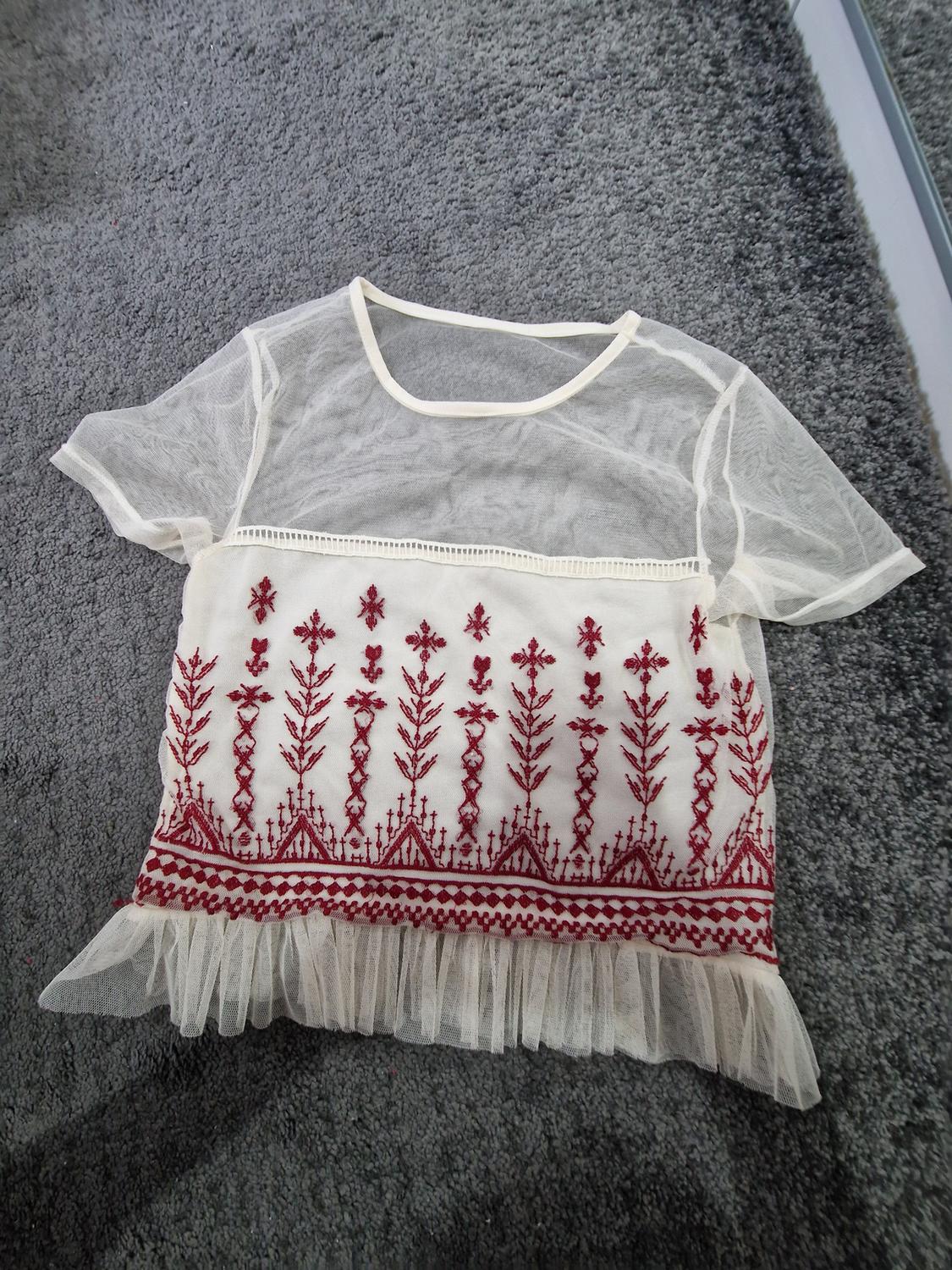 Cream-coloured semi-sheer short-sleeved top with red embroidery and a ruffled hem, on a grey rug.