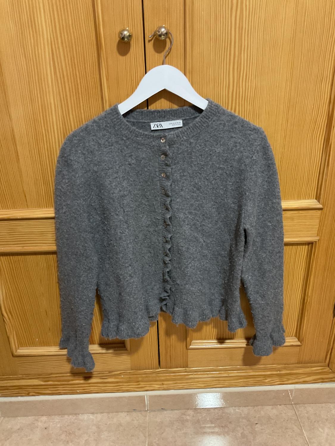 Grey wool cardigan with buttons and scalloped edges, hanging on a white hanger in front of a light wood wardrobe.