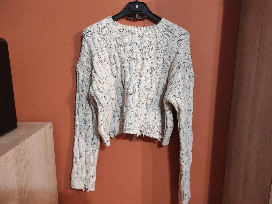 Chunky cream knitted jumper hanging on a black hanger against an orange wall. Light wood furniture in the background.