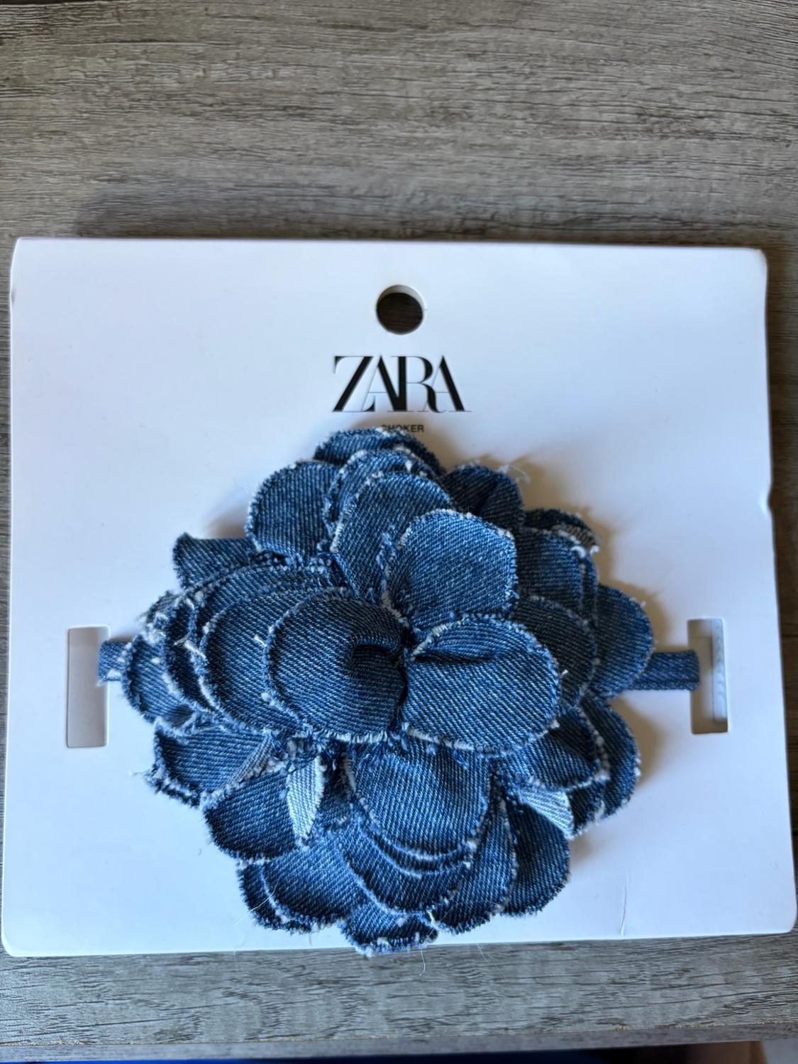 Blue denim flower choker with frayed edges on a white label bearing the text ZARA CHOKER, on a wooden surface.