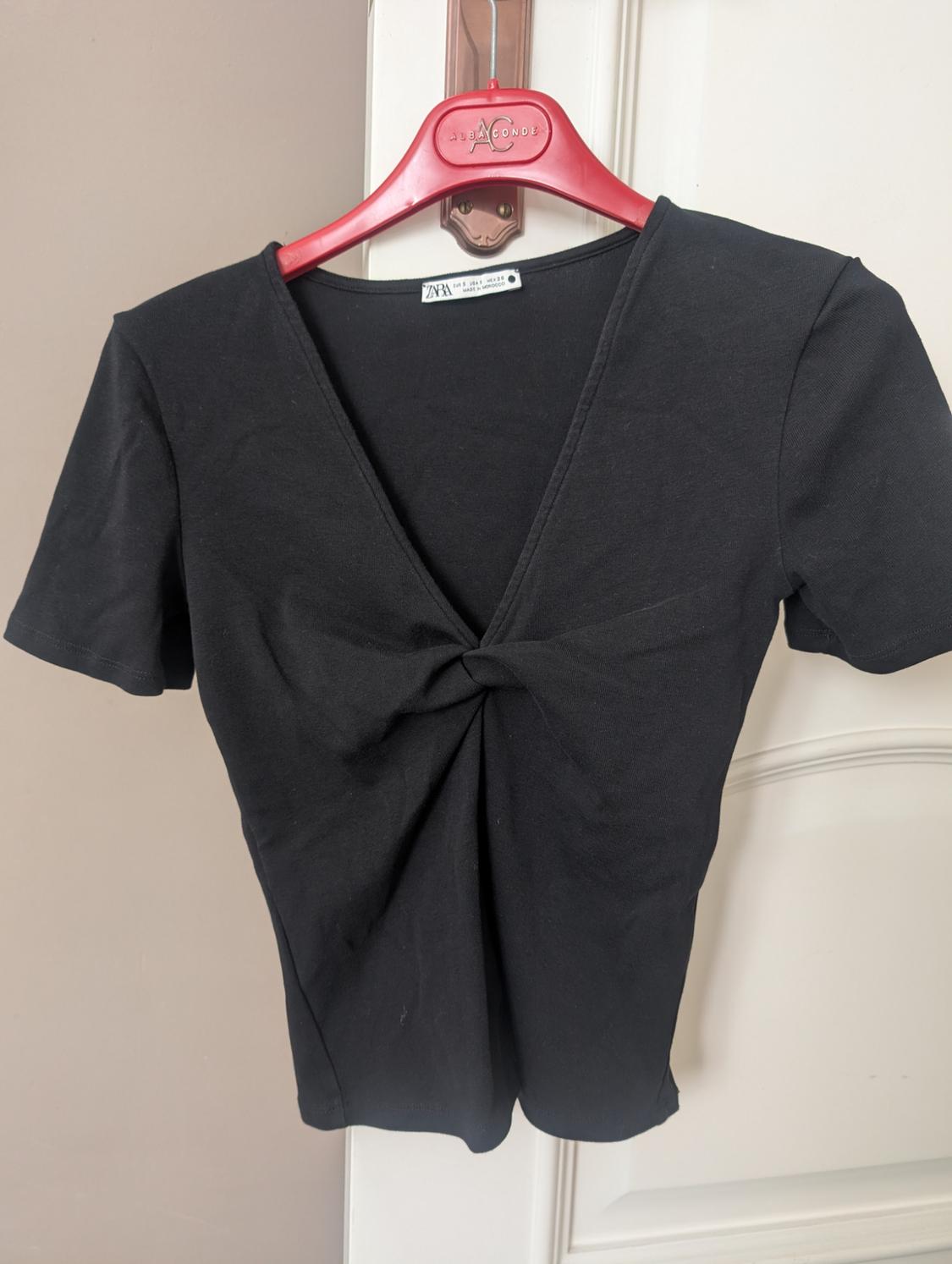 Black T-shirt with front knot, hanging on a red hanger with "Joa Conde" text, over a white door.